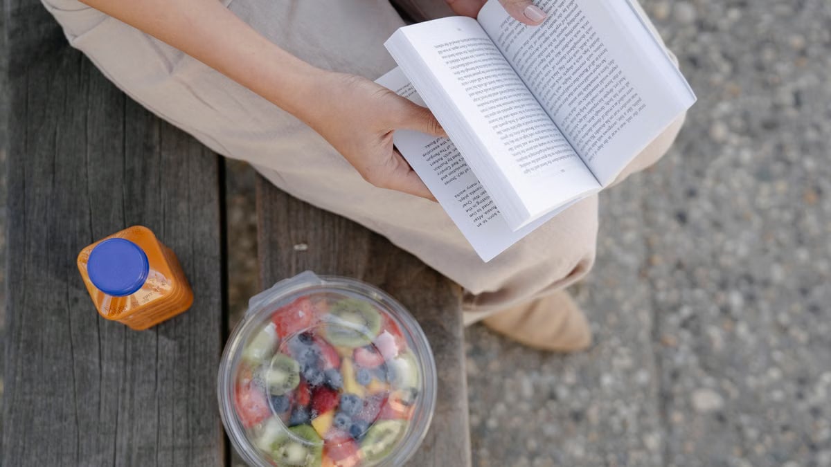 From above of crop unrecognizable female reading textbook on street bench with refreshing drink and fruit salad in takeaway container